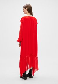 Vestido de chiffon rojo con mangas largas, dobladillo asimétrico y acentos con volantes. Combinado con botas negras de tobillo. La vista desde atrás muestra una silueta fluida.