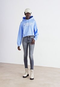 Haut cropped bleu clair avec logo blanc, jean skinny gris, et bottes de combat crème avec semelle noire, sur un fond blanc uni.