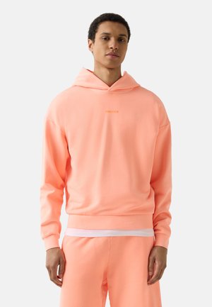 Młody mężczyzna ubrany w jasnoróżową bluzę z kapturem i pasujące spodnie dresowe, stojący na prostym białym tle.