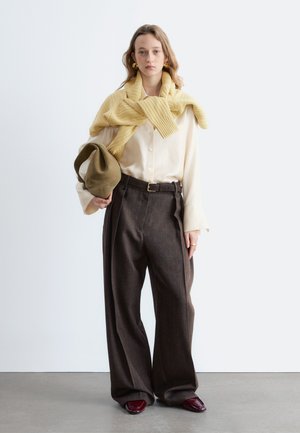 Giovane donna che indossa una camicetta color crema, un maglione giallo appoggiato sulle spalle, pantaloni larghi marrone scuro, scarpe rosse, che tiene una borsa verde oliva.