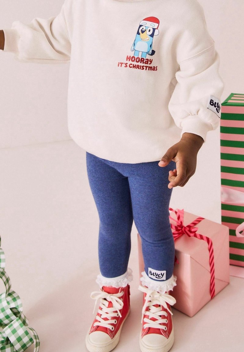 Enfant en leggings bleus, baskets rouges et sweat-shirt blanc avec dessin animé et texte "Hourra, c'est Noël", debout près de cadeaux de Noël emballés.