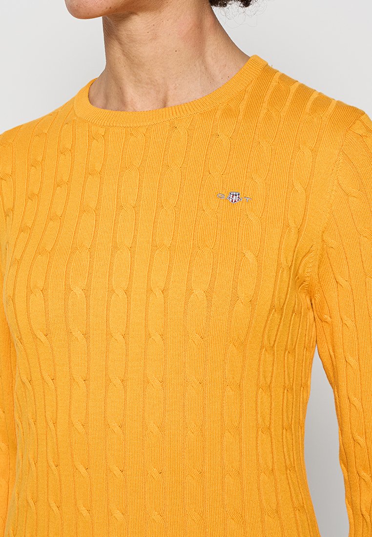 Żółty sweter dziany z wzorem w warkocze, okrągłym dekoltem i małym haftowanym logo na piersi. Tekstura wydaje się miękka i prążkowana.