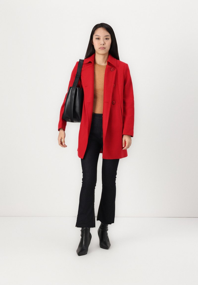Roter Wollmischungs-Mantel mit Reverskragen und doppelten Knöpfen, getragen über einem hellbraunen Pullover und dunklen Schlagjeans, kombiniert mit schwarzen Ankle Boots.