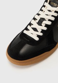 Black leather and suede sneaker with cream laces, featuring a rubber gum sole and triangular tread pattern. Coach logo in gold on the side.
