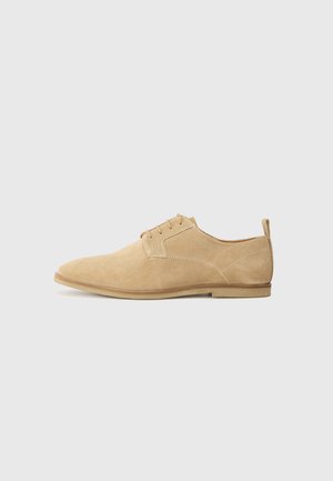 Chaussure Oxford pour homme en daim beige avec fermeture à lacets et petit talon, vue de profil sur un fond gris clair uni.