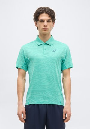 Jeune homme debout de face, portant un polo à motifs turquoise clair et un short bleu marine foncé sur un fond blanc uni.