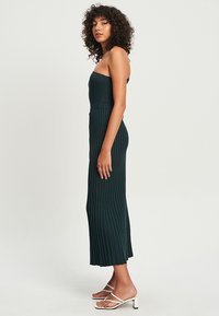 Langes, dunkelgrünes, geripptes Kleid mit einem taillierten, trägerlosen Oberteil und einem plissierten Rock, kombiniert mit weißen Sandalen mit Riemchen.