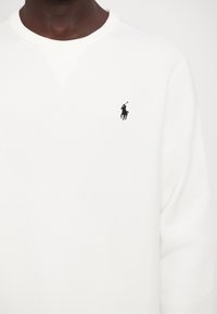 Personne portant un sweat-shirt blanc à col rond avec un petit logo noir de joueur de polo brodé sur la poitrine gauche.