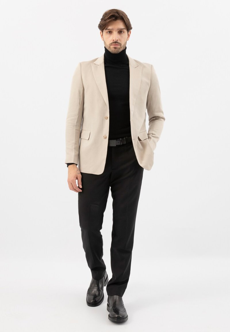 Uomo con blazer beige, dolcevita nero, pantaloni neri e scarpe nere in piedi con una mano in tasca su sfondo bianco.