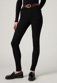 Femme portant un pantalon skinny noir, un haut à manches longues noir, une ceinture marron et des mocassins en cuir noir, debout devant un fond uni.
