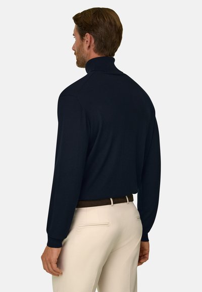 Homme portant un pull à col roulé bleu marine et un pantalon beige avec une ceinture marron, de dos, sur un fond clair uni.
