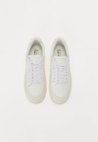 White sneakers with perforated design, contrasting beige accents, flat white laces, and a round toe. Smooth leather texture on upper.