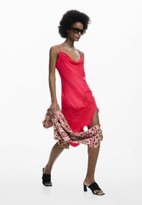 Vestido de satén rojo con escote redondeado y bordado floral en el dobladillo. La modelo sostiene un chal estampado y lleva mules negros de tacón y gafas de sol.