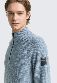 Pullover trui van textuur blauw breiwerk, met een hoge kraag en halve rits. Inclusief een zwart merklabel op de mouw.