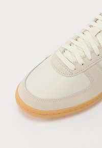 Beige színű sportcipő, textúrált velúrból és vászonból készült felsőrész, krémszínű fűzők és gumiból készült talp. Lekerekített orrdizájn, varrott díszítéssel.