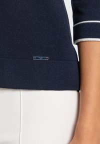 Haut tricoté bleu marine avec manches courtes ornées de rayures blanches, panneaux latéraux en accent, et un petit logo métallisé de la marque à l'ourlet.