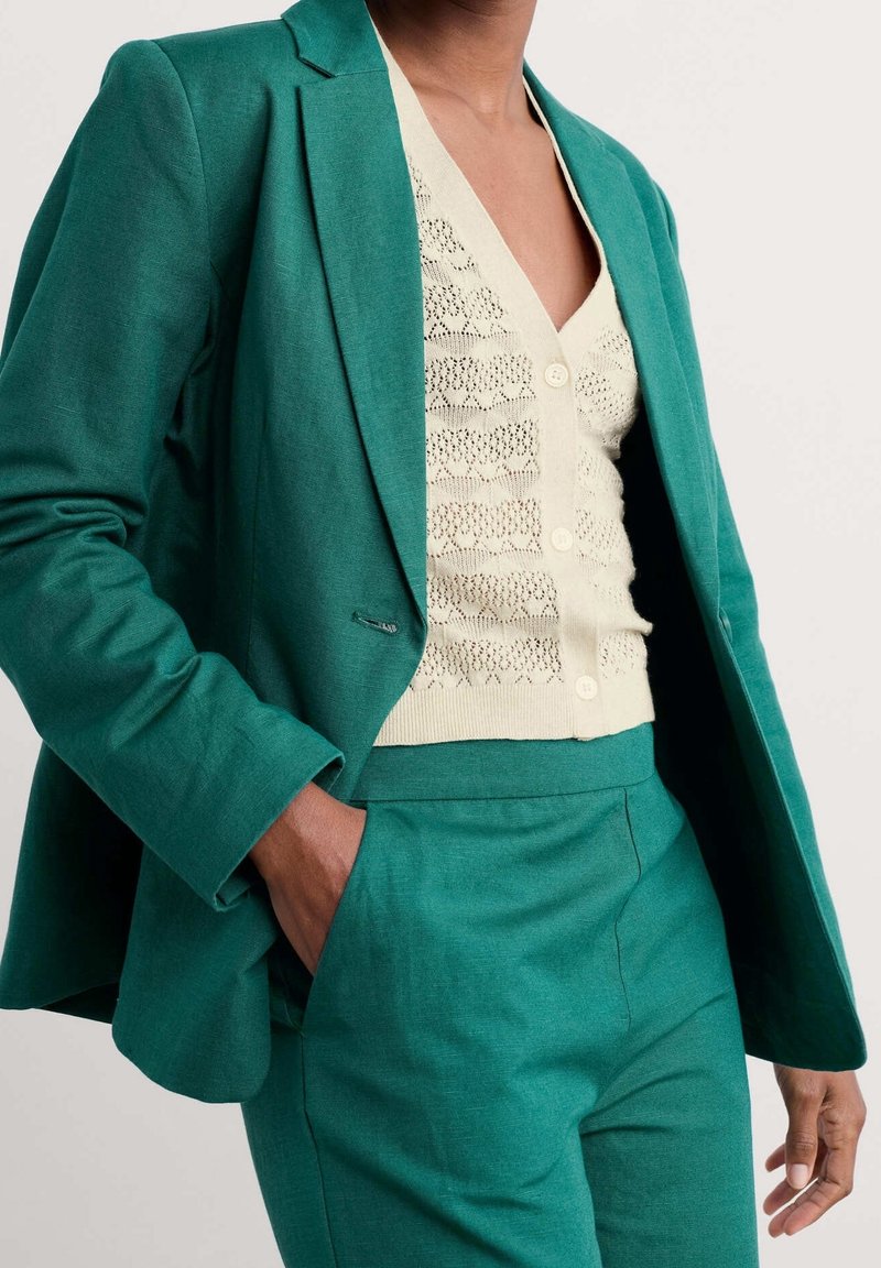 Blazer teal con taglio sartoriale, abbinato a un cardigan beige lavorato a maglia con bottoni e un motivo testurizzato. Pantaloni teal coordinati.
