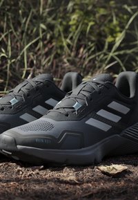 Σκούρα γκρι παπούτσια τρεξίματος Adidas με λευκές ρίγες σε χώμα, περιτριγυρισμένα από γρασίδι και φύλλα σε εξωτερικό χώρο.