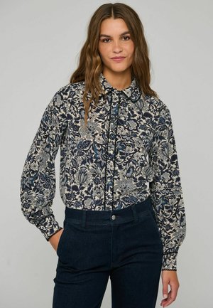 Frau mit langen, welligen Haaren, die eine langärmelige Bluse mit Blumenmuster trägt, in dunkle Hosen gesteckt, steht mit einer Hand in der Tasche.