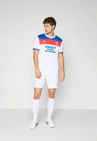 Bílý fotbalový dres s modrými a červenými akcenty, se znaky na hrudi. Krátké rukávy a šortky, doplněné bílými ponožkami a černými botami.