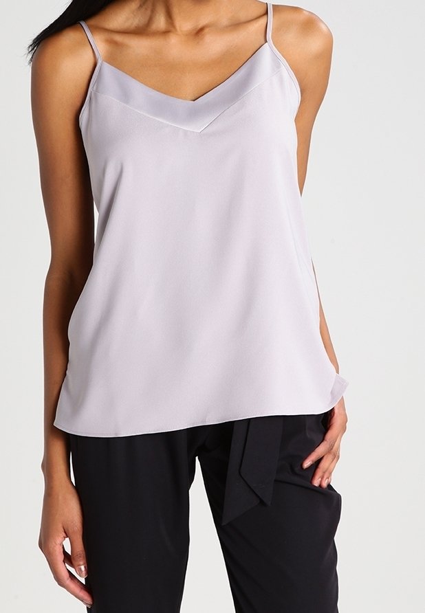 Kvinna iförd en ljus lavendelfärgad ärmlös topp med V-ringning och svarta högmidjade byxor med knytmidja, stående mot en enfärgad bakgrund.