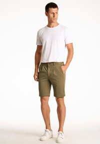 Vihreät oliivinväriset shortsit, joissa on sileä pinta, etutaskut ja nappisuljenta. Yhdistetty yksinkertaiseen valkoiseen t-paitaan ja valkoisiin lenkkareihin.