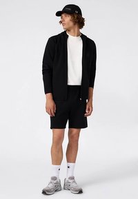 Chaqueta negra con cremallera, camiseta blanca, pantalones cortos negros, zapatillas grises y calcetines blancos. También lleva una gorra negra. Diseño sencillo y deportivo.