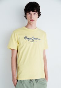 Geltona medvilninė marškinė su apvaliu kaklu, pasižyminti tamsiai mėlynu "Pepe Jeans London" logotipu įrašytu kursyvu per krūtinę. Paprastas dizainas.