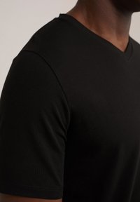 Tricou negru cu mânecă scurtă și decolteu în V, cu o textură netedă. Țesătura pare ușoară, se potrivește bine pe corp și are un finisaj curat la cusături.