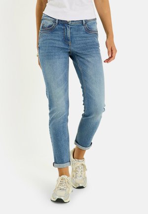 Persona que lleva vaqueros azules remangados y zapatillas blancas, de pie sobre un fondo claro y liso con torso inferior y piernas visibles.