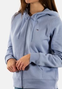 Felpa con cappuccio azzurro chiaro con cerniera, tasca frontale, coulisse regolabili, polsini a coste e piccolo logo sul petto. Tessuto morbido al tatto.