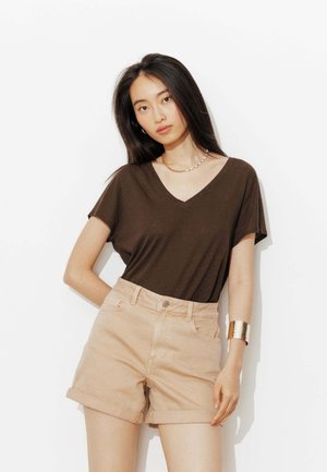 Femme aux cheveux longs et foncés portant un T-shirt marron à col en V, un short beige retroussé, un collier en perles et chaîne d'or, et un large bracelet en or.