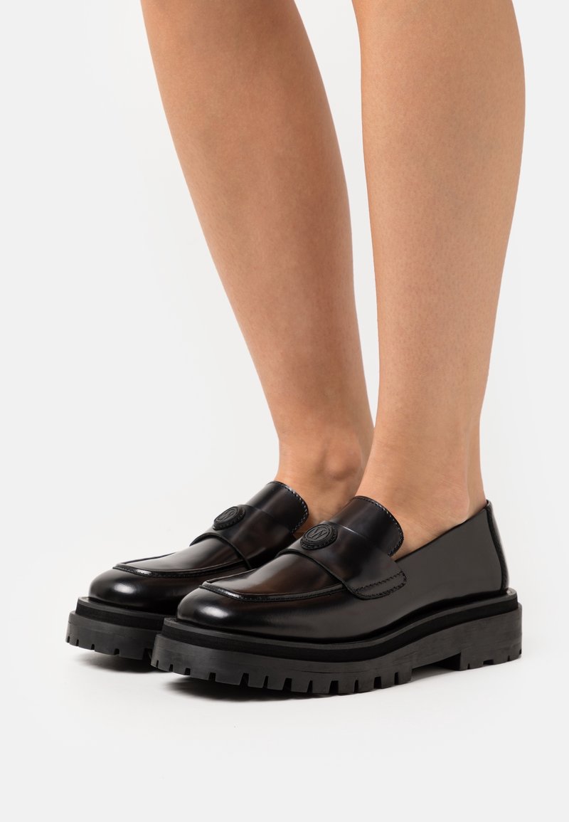 Mocassins en cuir noir avec une bande large et un détail de logo, dotés d'une semelle en caoutchouc noir épaisse et texturée, conçus pour un style à enfiler.