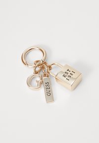 Porte-clés en or avec un pendentif en forme de "G", une étiquette rectangulaire gravée "GUESS" et un cadenas gravé "EST 1981" sur un fond blanc.