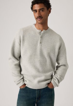 Uomo con capelli ricci e baffi che indossa un maglione grigio chiaro abbottonato e jeans blu con le mani in tasca su uno sfondo semplice.