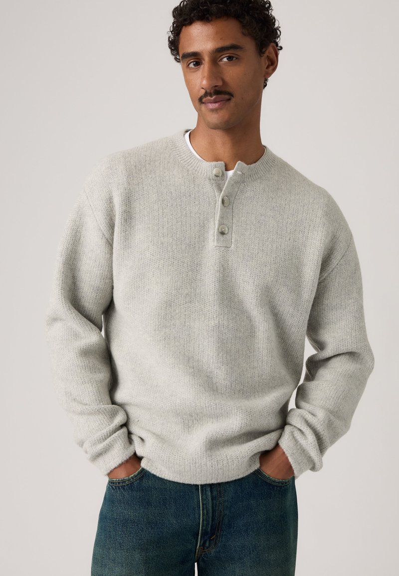 Homme aux cheveux bouclés et moustache portant un pull gris clair boutonné et un jean bleu, les mains dans les poches, sur un fond uni.