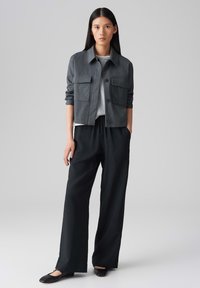 Grijs kort jack met twee voorzakken, gedragen over een lichtgrijze shirt. Het model heeft zwarte wijde broek en platte zwarte schoenen aan.