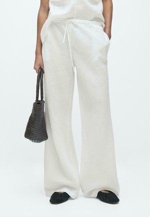 Personne portant un pantalon large en lin blanc avec une taille à cordon, des chaussures plates noires, et tenant un sac à main tissé marron foncé dans la main gauche.