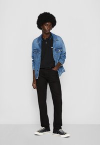 Afro soenguga ja mees, kellel on seljas sinine teksajakk, must polo, mustad teksad ja mustad tennised valgete taldadega, seismas valge taustaga.