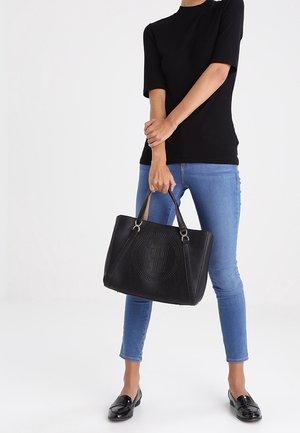 Czarna torba na ramię ze skóry z perforowanym wzorem w kształcie kół, podwójnymi uchwytami i usztywnionym kształtem. Modelka nosi niebieskie dżinsy i czarną bluzkę.