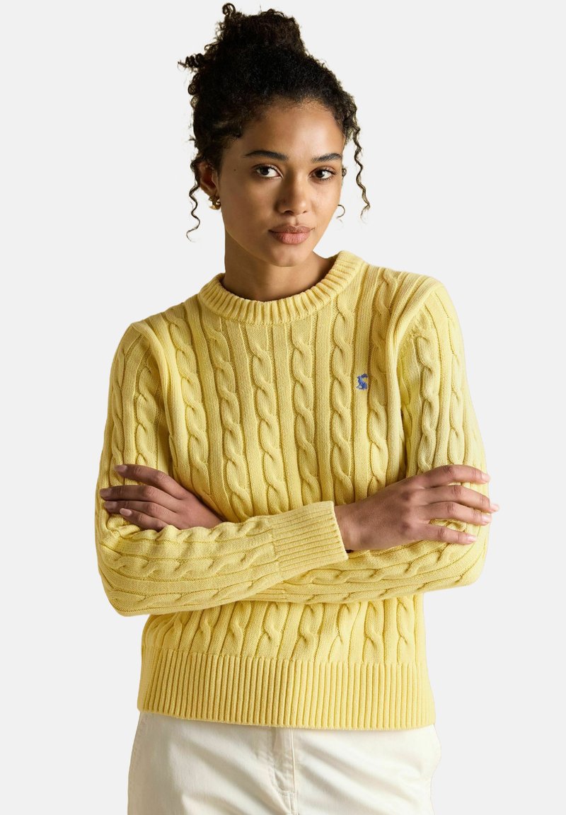 Moteris su garbanotais plaukais sukryžiuotais rankomis, vilkinti geltoną megztinį su pynėmis ir kreminės spalvos kelnes, prieš baltą foną.