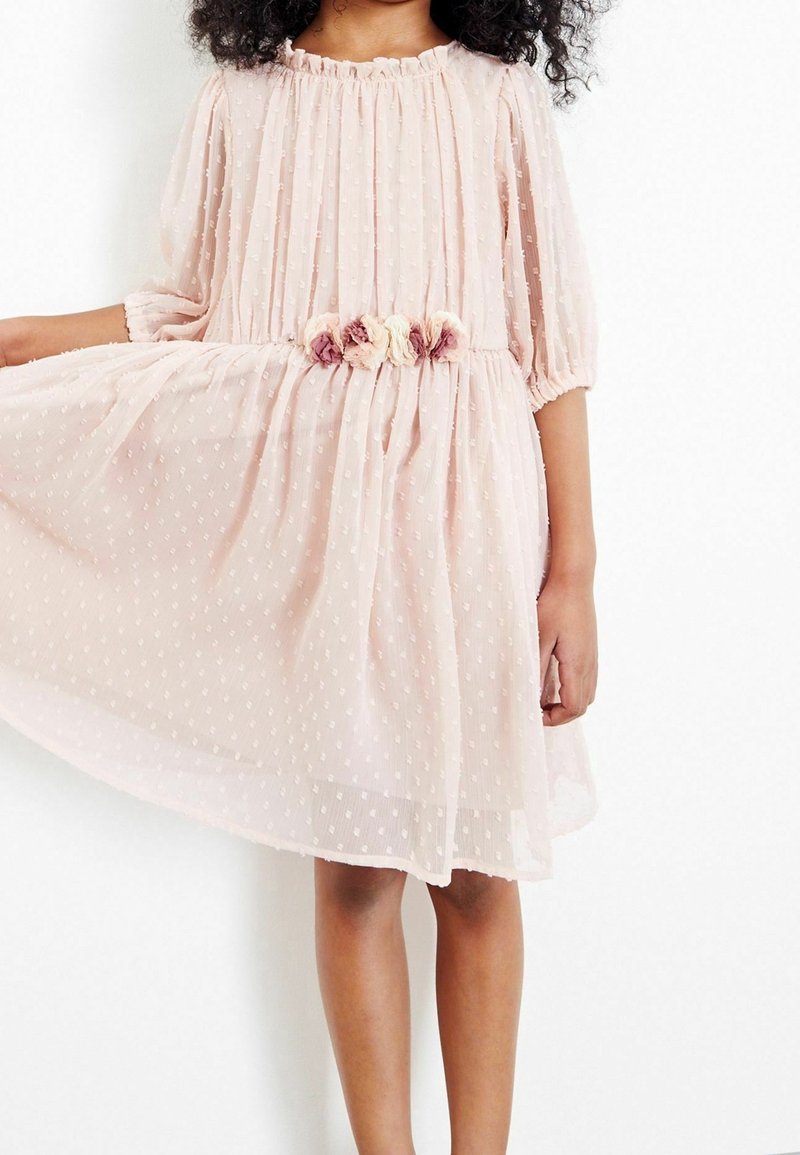 Robe rose clair à manches courtes en tulle à pois ; elle présente une taille froncée avec un accent floral aux couleurs douces.