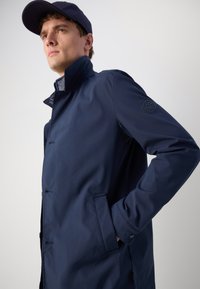 Giacca impermeabile navy con colletto alto, chiusura a bottoni, tasche laterali e un logo sulla manica. Texture liscia e design moderno.