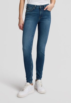 Person trägt blaue Slim-Fit-Jeans und weiße Sneaker, steht mit einer Hand in der Tasche vor einem einfarbigen Hintergrund.