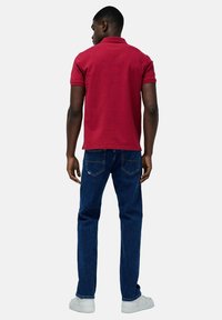 Polo shirt in cotone rosso con maniche corte, abbinato a jeans blu scuro. L'outfit è completato da sneakers bianche. Visto da dietro.