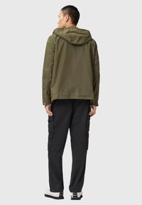 Veste à capuche vert olive avec une coupe décontractée, en matériau léger, et accents jaunes distinctifs sur les manches. Associée à un pantalon cargo noir.