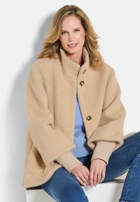 Beige gebreide jas met een hoge kraag, drie grote knopen en brede mouwen, gedragen over een blauwe top en gecombineerd met jeans. Getextureerde stof.