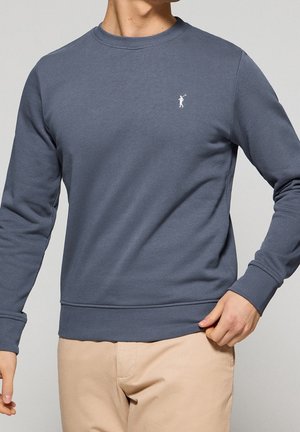 Mann trägt ein blaues Sweatshirt mit Rundhalsausschnitt und kleinem weißem Polo-Spieler-Logo auf der Brust und beige Hose, steht vor einem einfachen Hintergrund.