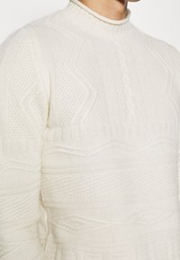 Pull en maille couleur crème avec un col haut. Présente des motifs texturés, notamment des tresses et des formes géométriques sur toute sa surface.