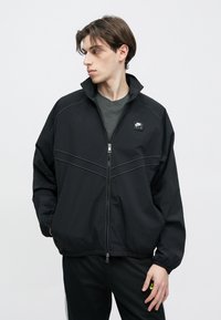 Μαύρο ελαφρύ μπουφάν με φερμουάρ μπροστά, ψηλό γιακά, μανίκια με μανσέτες και διακριτικές ραφές. Διαθέτει μικρό λογότυπο στο στήθος.